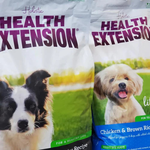 Two bags of Holistic Health Extension dog food show close-up images of a black and white dog and a fluffy beige dog, set against a grassy background.