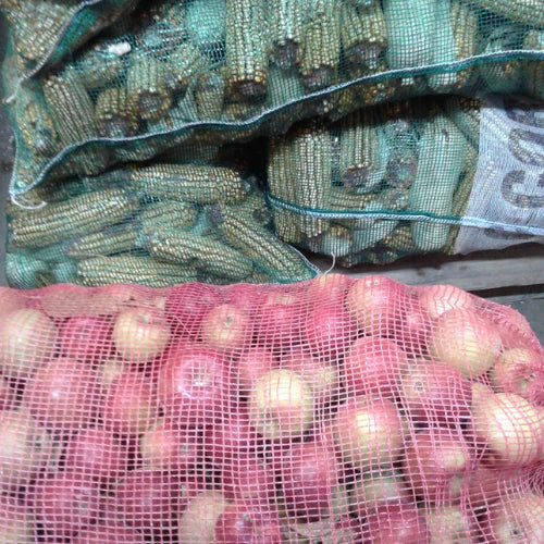 Bags of corn and onions in mesh sacks are stacked, showcasing fresh produce. The colors are natural, conveying a rustic, market-like feel.